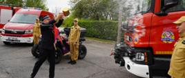 Kapelan Diecezjalny Strażaków w uroczystym geście polewa wodą święconą z wiaderka, przód nowego wozu ratowniczo-gaśniczego. W tle strażacy kierowcy stojący przy nowych pojazdach pożarniczych ubraniu w mundury służbowe. 