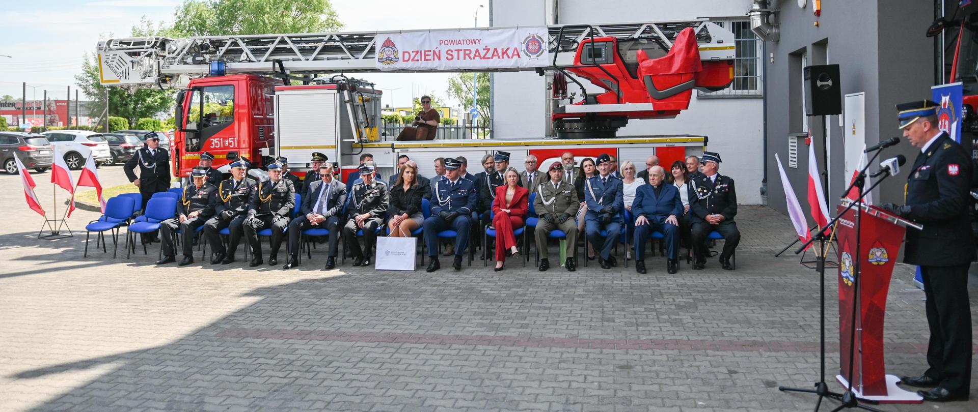 Goście siedzą na tle pojazdu strażackiego w kolorze czerwonym, jeden strażak przemawia.