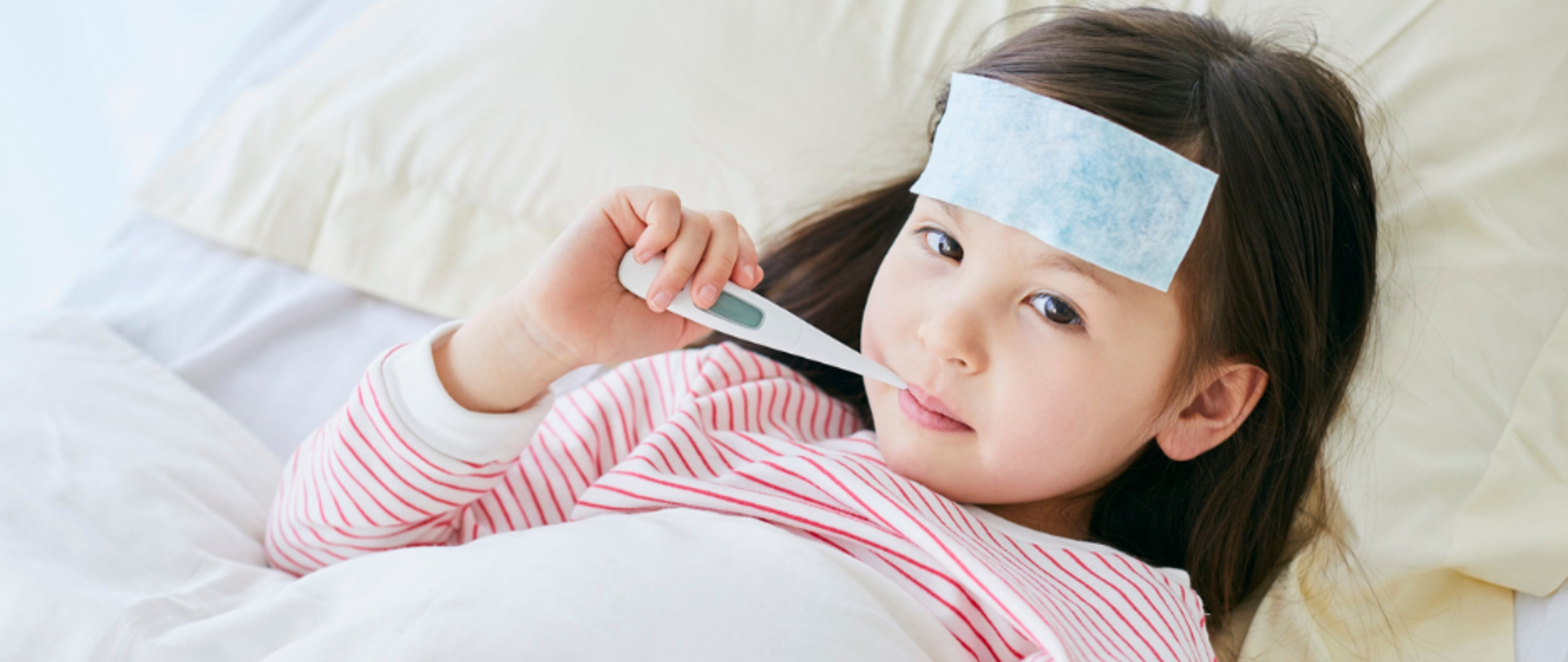 Chora dziewczynka leży w łóżku pod kołdrą z termometrem w ustach.