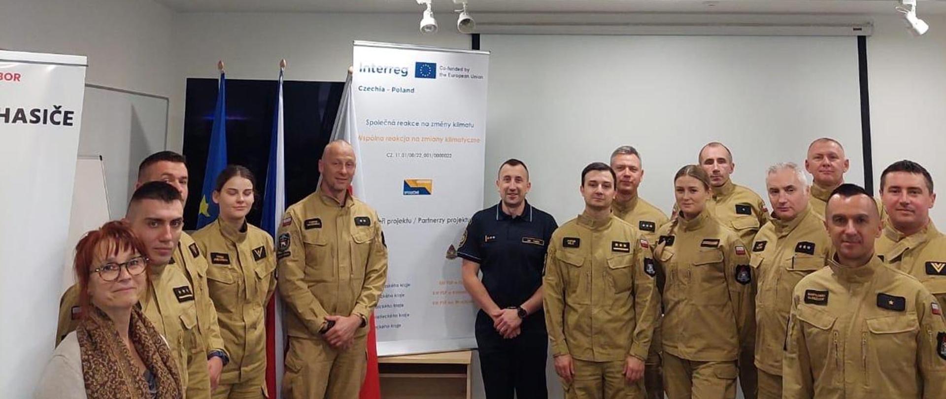 Grupa strażaków ubranych w ubrania służbowe w kolorze piaskowym oraz kilku cywili pozuje do zdjęcia na tle baneru promującego porogram Interreg