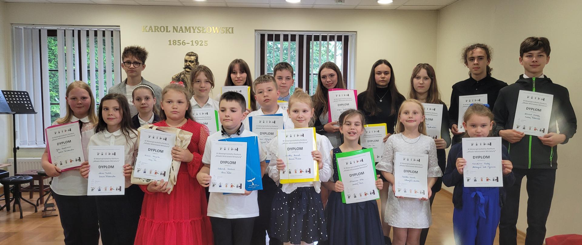 Ustawione w trzech rzędach dzieci trzymają w rękach białe kartki - Dyplomy. Wszystkich uczestników jest 19 osób: 13 dziewczynek i 6 chłopców. Dzieci uśmiechnięte, w strojach galowych. W dali widać dwa okna, stojący z boku pulpit. Na ścianie postument z napisem Karol Namysłowski 1856-1925. 