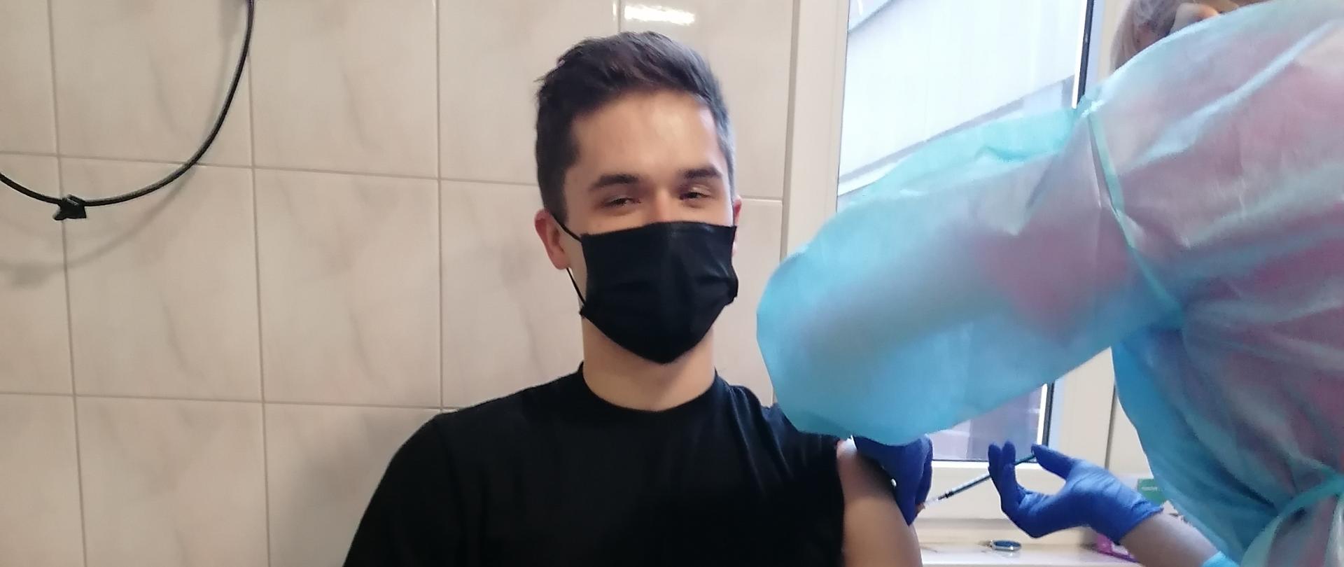 Zdjęcie przedstawia strażaka siedzącego, przyjmującego dawkę szczepienia przeciw COVID-19