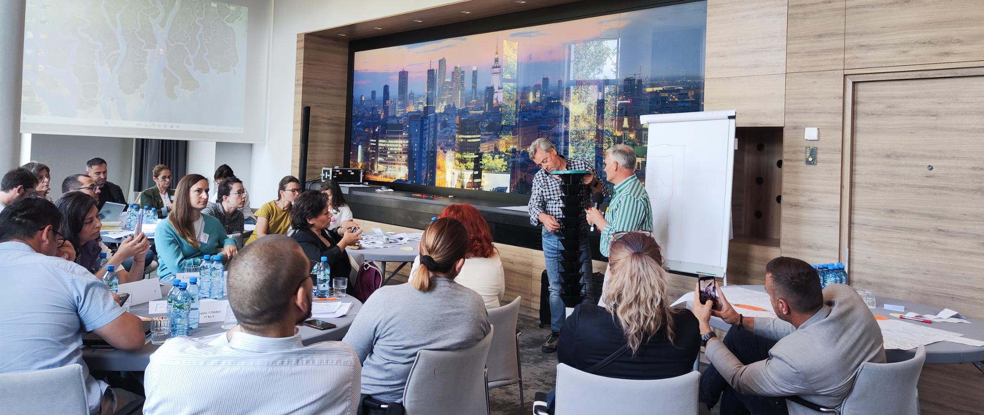 Dwóch mężczyzn prezentuje pułapkę do odłowu Monochamus. Pułapka składa się z kilku czarnych lejków przykrytych zieloną pokrywką. Na sali przy okrągłych stołach siedzą uczestnicy szkolenia. W tle tapeta z panoramą miasta Warszawa.