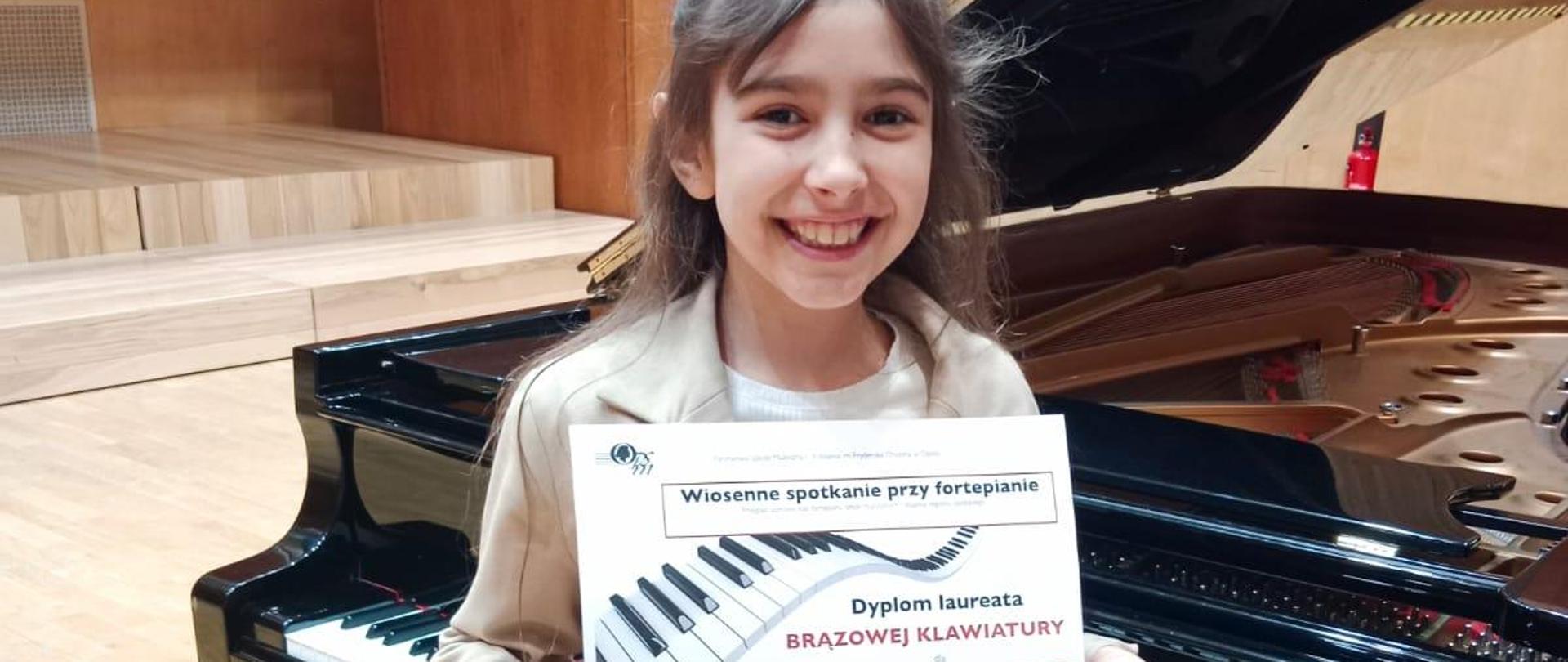 Zdjęcie szeroko uśmiechniętej dziewczynki - Igi Węglowskiej z dyplomem laureata brązowej klawiatury przed sobą. Dziewczynka ma ciemne włosy i beżowe ubranie stoi na scenie na tle otwartego fortepianu. 