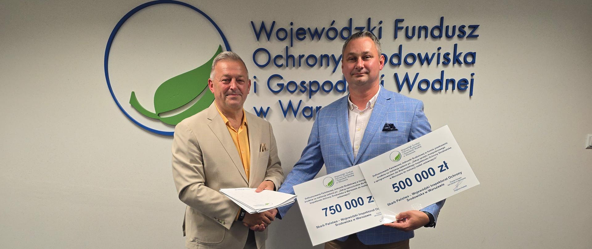 Dwóch mężczyzn pozuje do pamiątkowego zdjęcia na tle loga Wojewódzkiego Funduszu Ochrony Środowiska i Gospodarki Wodnej w Warszawie. Od lewej stoją: Prezes Zarządu WFOŚiGW w Warszawie Zbigniew Deptuła i Mazowiecki Wojewódzki Inspektor Ochrony Środowiska Krzysztof Cząstkiewicz.
