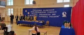 Na zdjęciu widać komisje egzaminacyjną i dzieci siedzące w ławkach. Komendant Powiatowy PSP wita wszystkich przybyłych. Po prawej stronie stoją puchary i pozostałe nagrody. Na wprost stoi niebieski baner z nazwą wydarzenia.