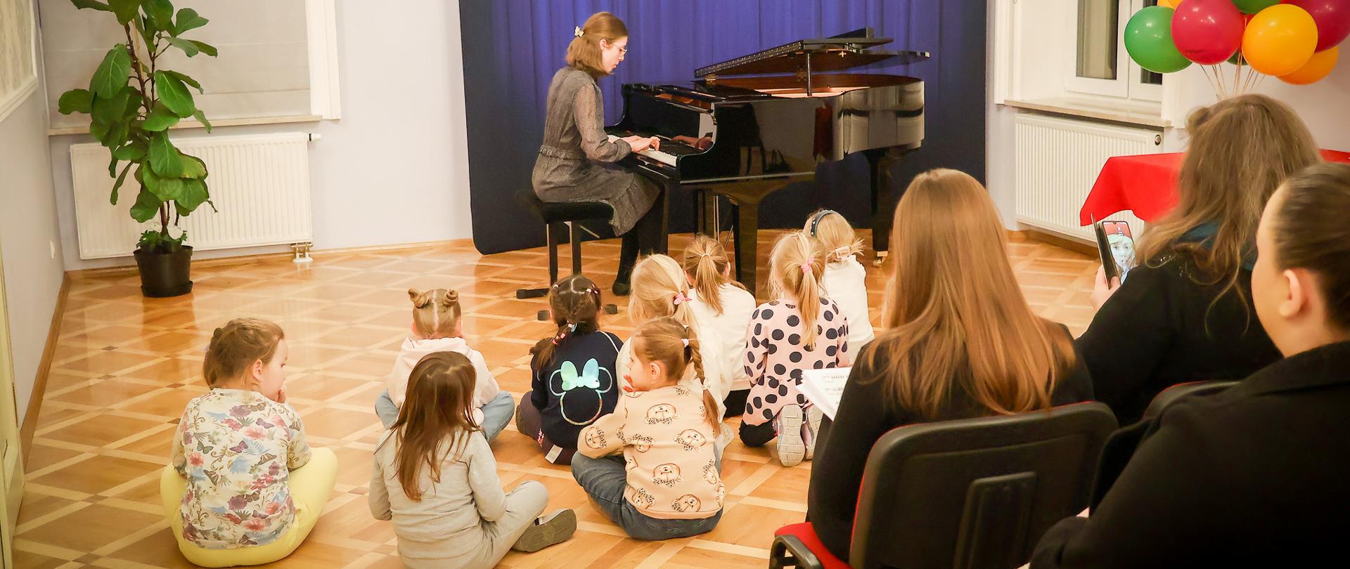 Dziewczynka grająca na fortepianie podczas występu w szkole muzycznej, siedząca na scenie przed grupą małych dzieci siedzących na podłodze i oglądających koncert; w tle widoczna ścianka z logo szkoły, a po prawej stronie kolorowe balony i dorośli widzowie.