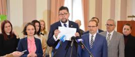 Wiceminister Szeptycki stoi i mówi do mikrofonu na stojaku, w rękach trzyma dokument, dookoła niego duża grupa osób, w tle zasłonięte okna.