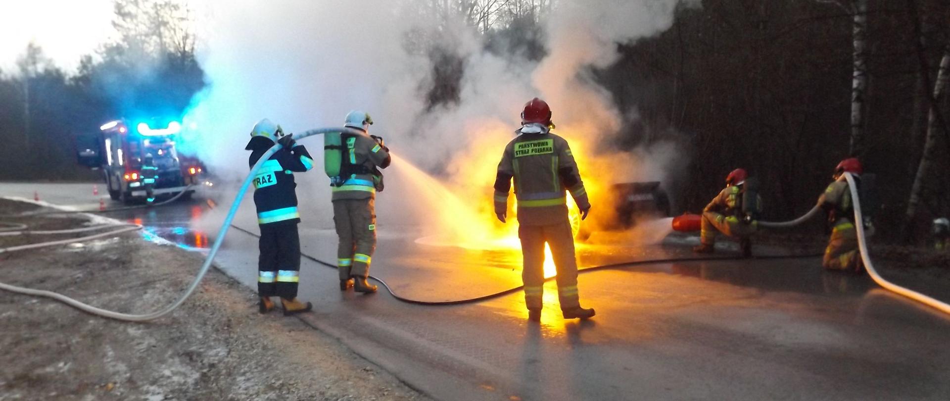 Na zdjęciu znajdują się strażacy gaszący pożar samochodu. Samochód znajduje się na drodze przy lesie - w tle samochód strażacki na sygnałach świetlnych.