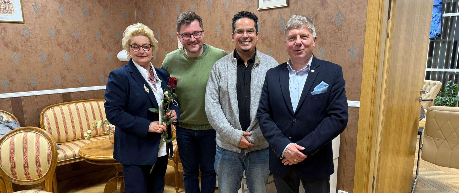 Na zdjęciu grupa osób. Po lewej stronie kobieta blondynka w okularach w czarnym garniturze, która trzyma czerwoną róże. Obok niej trzech mężczyzn. Pierwszy w zielonym sweterku i czarnych spodniach, dalej mężczyzna w czarnej koszuli, jeansowych spodniach i szarym sweterku. Na końcu mężczyzna w jasnej koszuli i granatowym garniturze. Tło obrazka w odcieniach brązu. Na ścianach pomieszczenia dekoracyjna tapeta.