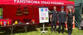 Trzech funkcjonariuszy Państwowej Straży Pożarnej – dwóch mężczyzn i jedna kobieta – pozuje przed czerwonym namiotem promocyjnym z logo PSP oraz białym napisem Państwowa Straż Pożarna podczas wydarzenia plenerowego. Obok znajdują się elementy wyposażenia strażackiego, hełmy, torby ratownicze i tablica informacyjna pod namiotem.