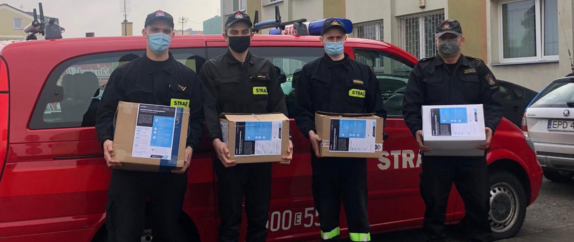 Czterej funkcjonariusze stoją na tle samochody osobowego strażackiego. Każdy z nich trzyma kartonowe pudełko z naklejoną ulotką,
