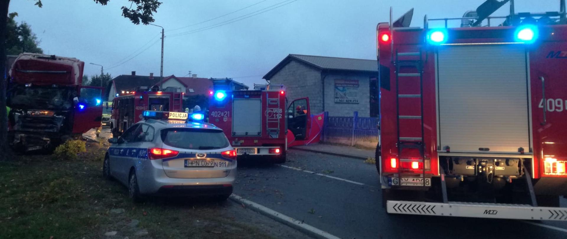 Zdjęcie przedstawia 5 samochodów
3 z nich to wozy bojowe straży pożarnej,
które mają włączoną sygnalizację świetlną stoją na ulicy
po lewej stronie widaż duże drzewo a przed nim samochód ciężarowy,
który uderzył w to drzewo jego przód jest uszkodzony,
na pierwszym planie stoi radiowóz policyjny który również ma włączoną sygnalizację świetlną,
w oddali widać zabudowania, pomiędzy wozami strażackimi jest rozstawiony parawan nad którym pochylają się strażacy.
