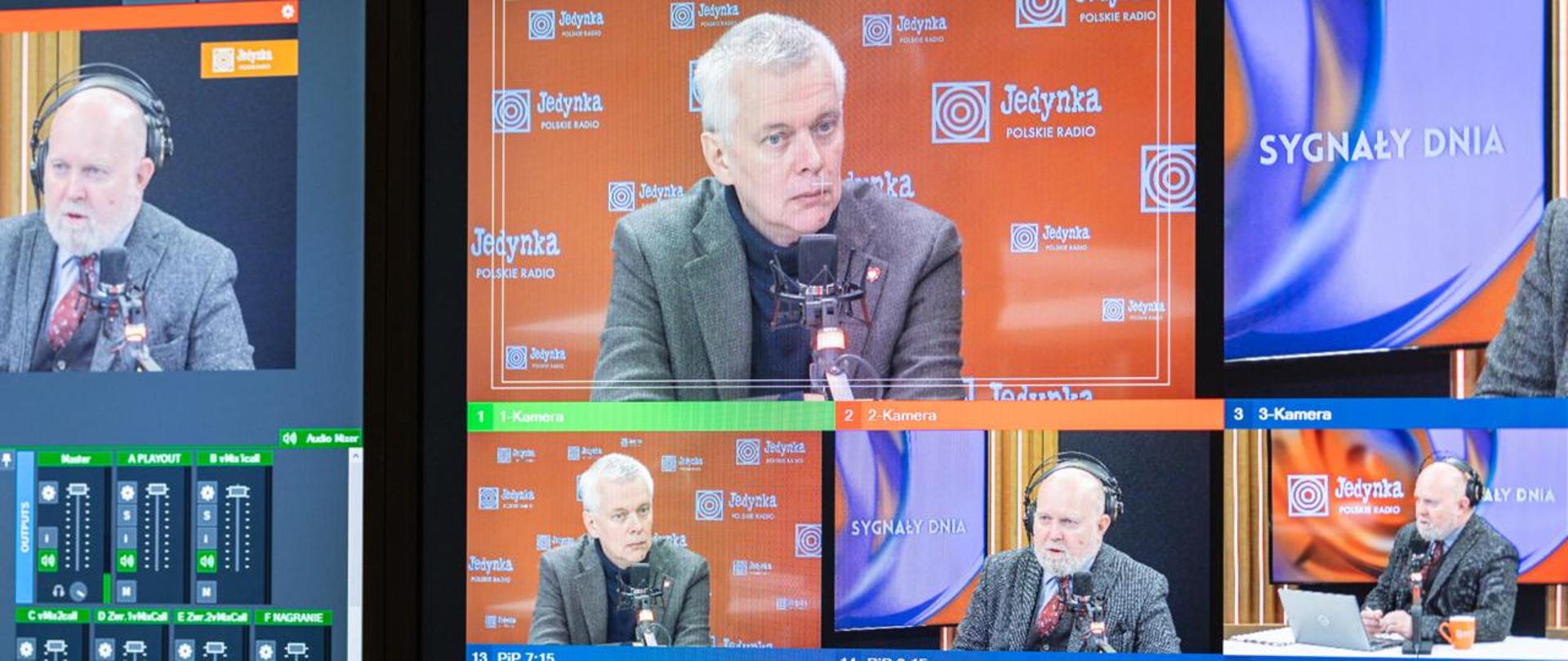 Stanowisko realizacyjne w studiu radiowym. Na ścianie wiszą dwa duże monitory. Na lewym ekranie widoczny jest interfejs programu do realizacji transmisji na żywo, z podglądem obrazu z kamer, listą źródeł sygnału oraz mikserem audio z suwakami i wskaźnikami poziomu dźwięku. Na prawym monitorze wyświetla się podgląd wielokamerowy audycji: w głównym oknie widać siwowłosego mężczyznę w szarej marynarce i czarnym golfie, siedzącego przy mikrofonie na tle pomarańczowej ściany z logotypami Polskiego Radia Jedynki. W pozostałych oknach widoczni są drugi mężczyzna w słuchawkach, siedzący przed mikrofonem, oraz szersze ujęcia studia z prowadzącymi przy stole. W dolnej części ekranu znajdują się dodatkowe podglądy kamer i plansze z nazwiskami. Na pierwszym planie, na drewnianym blacie, leżą konsoleta sterująca, joystick do obsługi kamer oraz inne urządzenia techniczne. Zdjęcie przedstawia zaplecze techniczne realizacji audycji radiowej z wykorzystaniem obrazu z kilku kamer.
