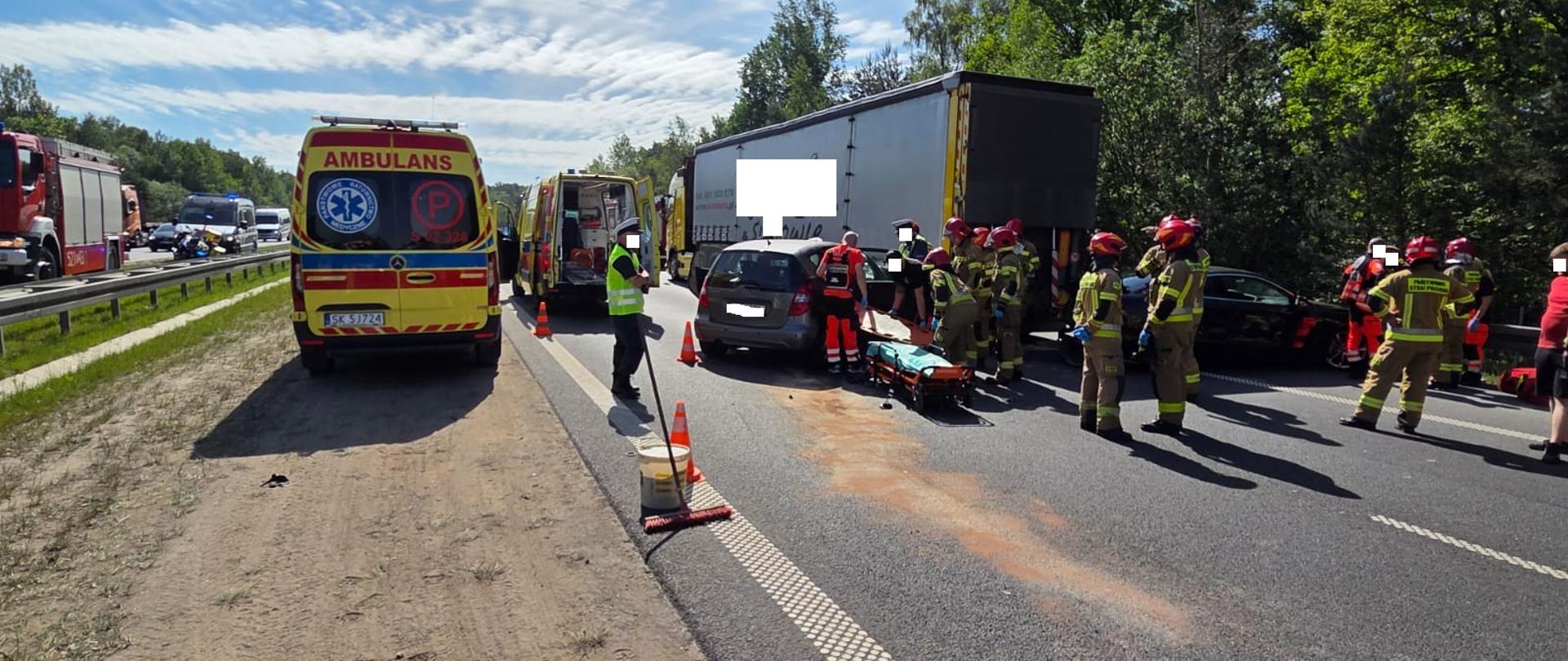 Zdjęcie przedstawia intensywną akcję ratunkową na zablokowanej drodze po wypadku z udziałem ciężarówki i samochodów osobowych, gdzie obok stojących ambulansów strażacy oraz ratownicy medyczni przygotowują nosze i udzielają pomocy poszkodowanym.