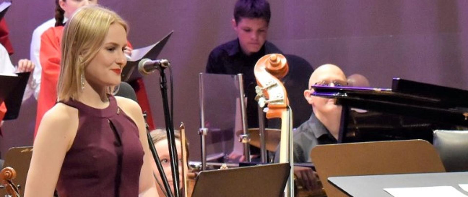 Zdjęcie kolorowe. Wokalistka w amarantowym stroju wieczorowym stoi przy mikrofonie. W tle pulpity, instrumenty i uczniowie orkiestry szkolnej.