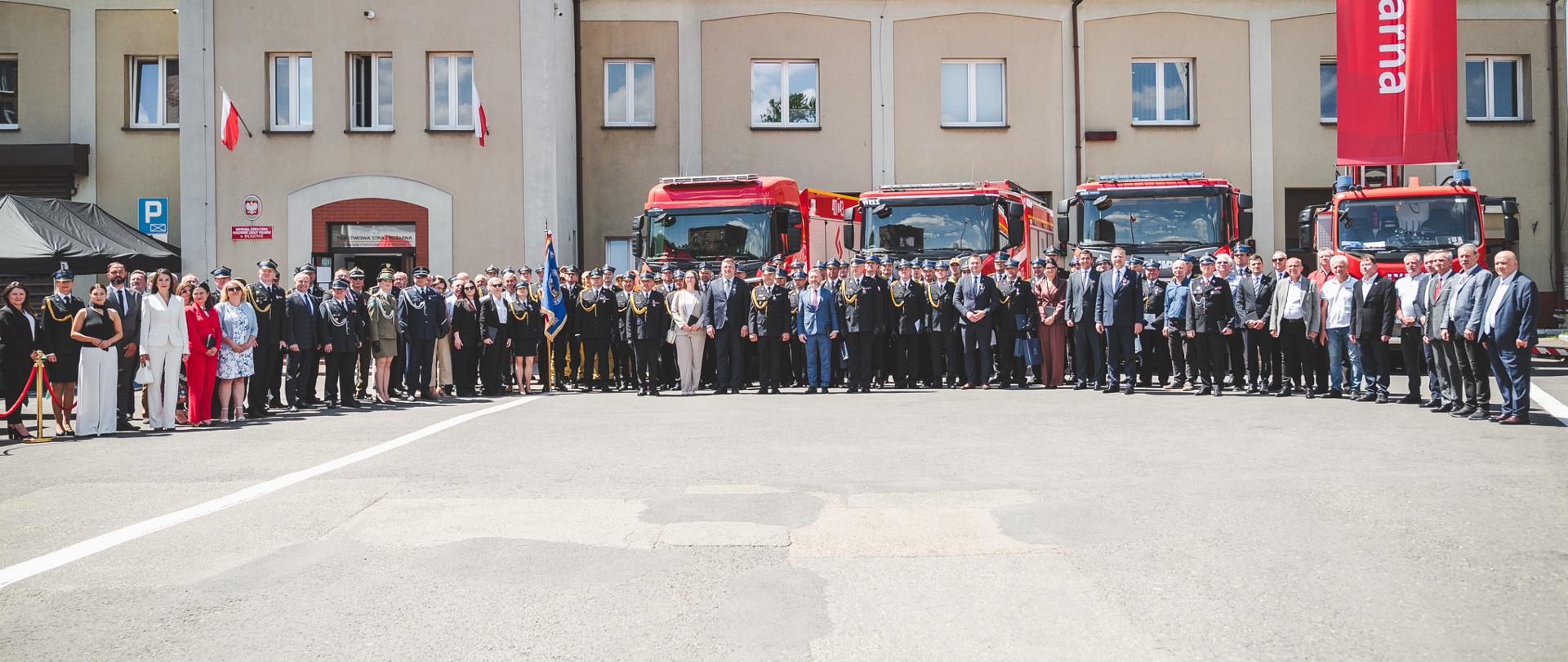 Szerokie ujęcie grupowego zdjęcia wykonanego przed budynkiem straży pożarnej z widocznym napisem „STRAŻ POŻARNA” na elewacji. Na placu stoi duża grupa osób – strażacy w mundurach galowych oraz osoby w garniturach i eleganckich strojach – ustawieni w jednym rzędzie do fotografii. W tle widoczne są czerwone wozy strażackie zaparkowane przed budynkiem oraz wysoki czerwony baner. Na dachu budynku znajdują się panele słoneczne, a na elewacji wiszą biało-czerwone flagi. Scena odbywa się na świeżym powietrzu w słoneczny dzień, a całość ma charakter oficjalnej uroczystości.