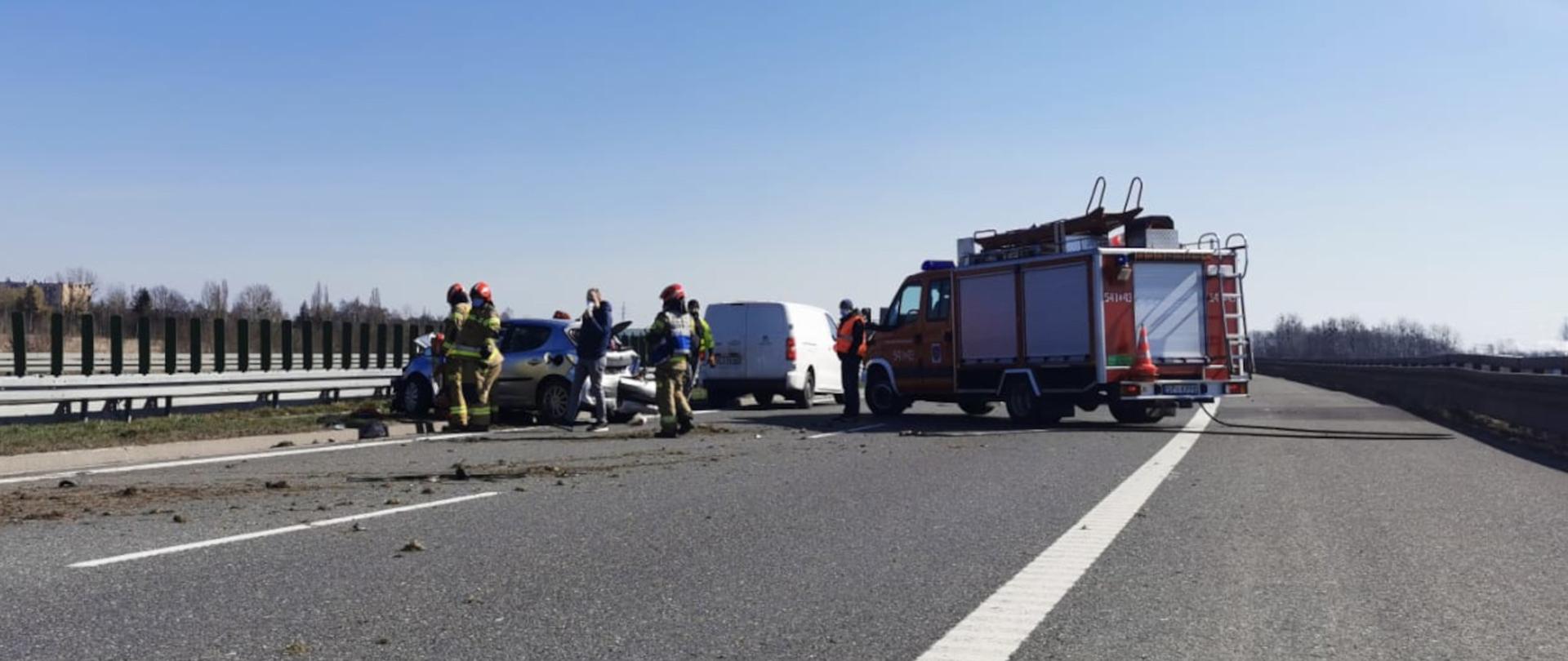 Zdjęcie przedstawia od lewej rozbity samochód a wokół niego strażaków ubraniach specjalnych. Następnie widać biały samochód dostawczy oraz lekki samochód ratownictwa technicznego.. Na jezdni widać rozrzuconą ziemię i ślady kół.