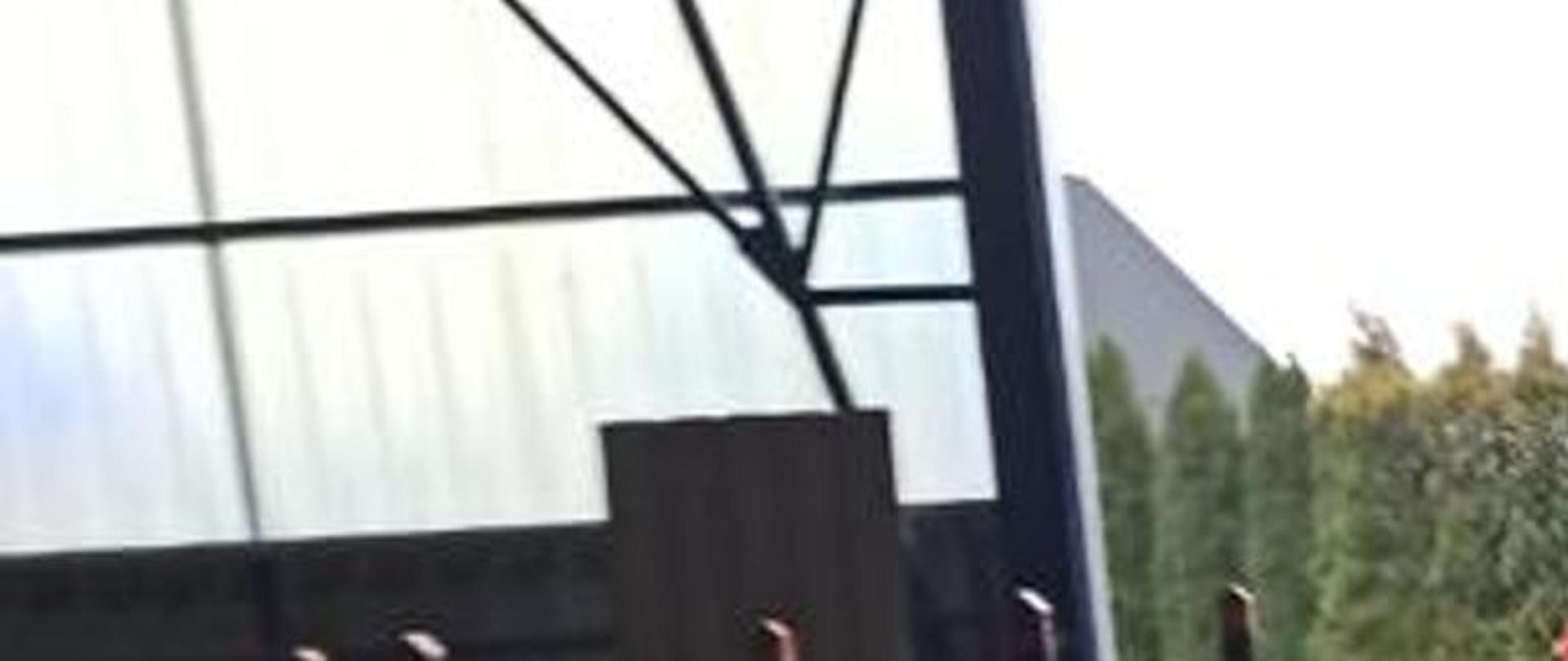 Zdjęcie wykonane wewnątrz budynku, w którym doszło do pożaru. Na pierwszym planie rozwinięty wąż od samochodu strażackiego, który znajduje się na dalszym planie, ustawiony bokiem. Przy pojeździe stoi strażak w ubraniu specjalnym.