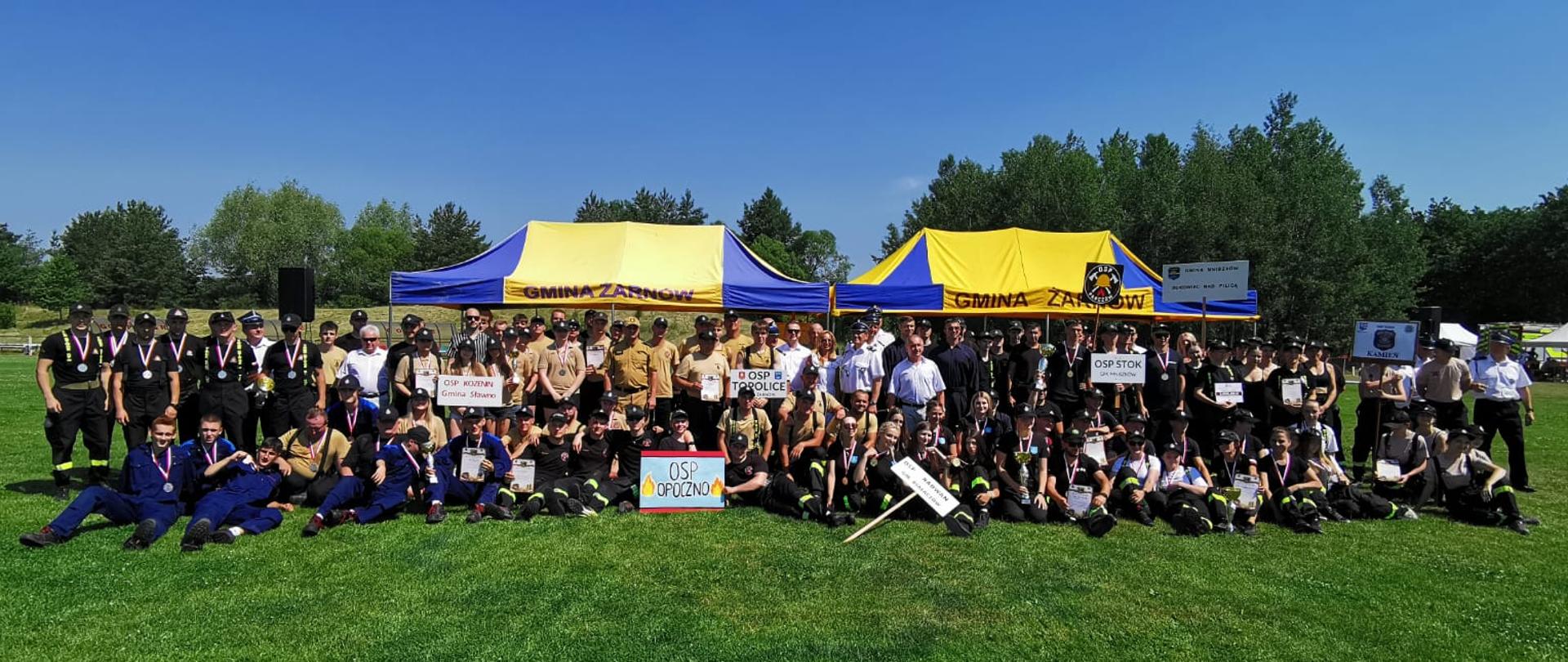 Wspólne zdjęcie grupowe na boisku trawiastym, zaraz po rozdaniu medali. Strażacy OSP z banerami nazw własnych jednostek OSP, nagrodzeni strażacy OSP z medalami, sędziowie oraz goście stojący po środku. W tle namioty z napisami Gmina Żarnów. 