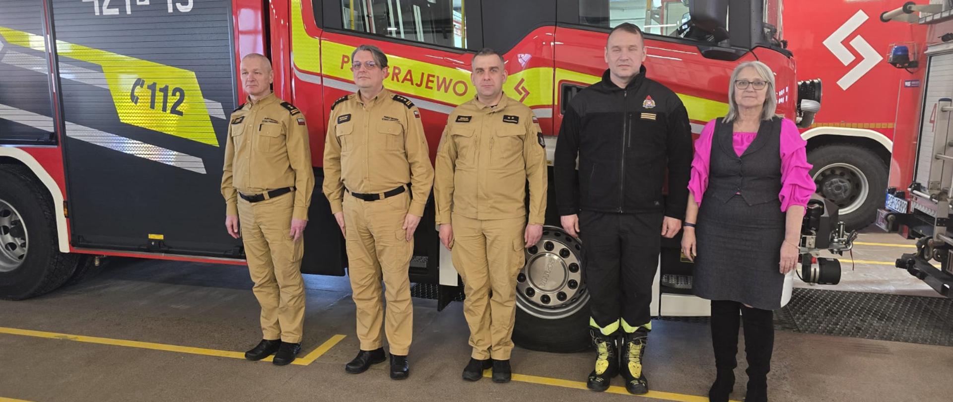 16 lutego 2026 r. w Komendzie Powiatowej Państwowej Straży Pożarnej w Grajewie odbyła się narada podsumowująca funkcjonowanie ochrony przeciwpożarowej na terenie powiatu grajewskiego w 2025 roku- strażacy ma czele z zastępcą podlaskiego komendanta wojewódzkiego PSP i pani wicestarosta na tle auta pożarniczego w garażu PSP