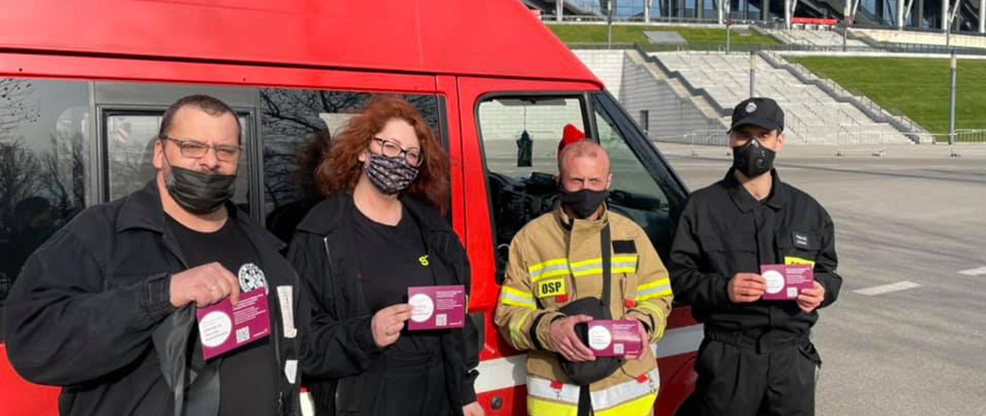 Pamiątkowe zdjęcie po wykonanym szczepieniu przeciw Covid-19. Na tle Stadionu Narodowego w Warszawie widać zaparkowany osobowy bus jednostki OSP. Przed samochodem, w rzędzie stoją trzej druhowie oraz druhna. W ręku trzymają potwierdzenie wykonania szczepienia. 