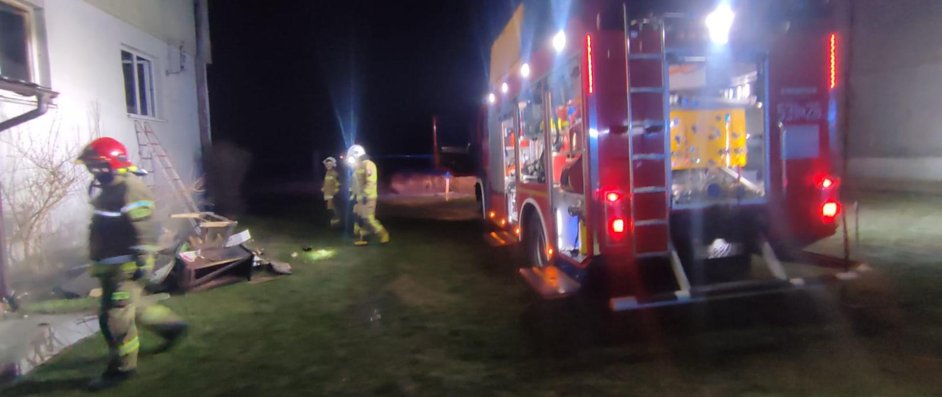 Po lewej budynek mieszkalny, po prawej samochód pożarniczy, pomiędzy strażacy