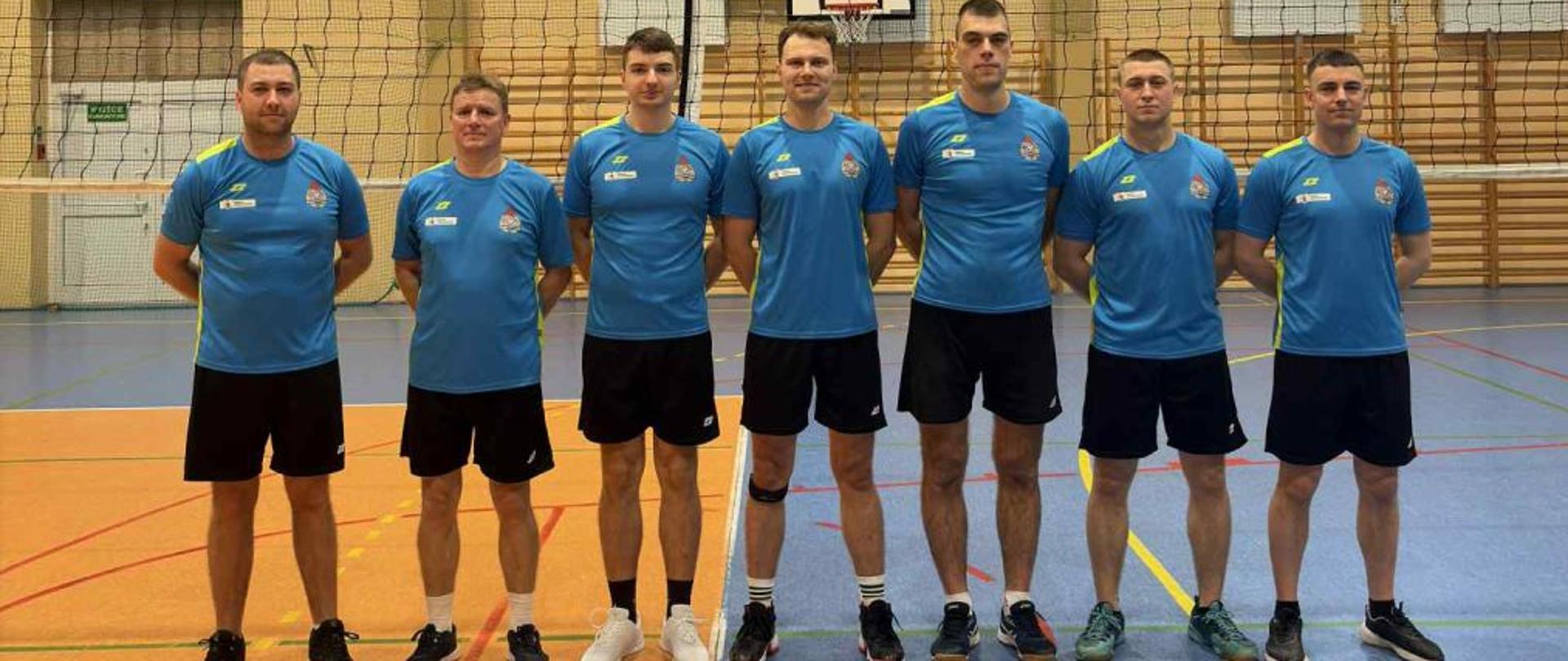 Na zdjęciu widoczna jest drużyna KP PSP Białogard ustawiona do wspólnej fotografii na hali sportowej. Zawodnicy ubrani są w jednolite stroje sportowe, co podkreśla zespołowy charakter udziału w turnieju. W tle widoczna jest siatka do piłki siatkowej oraz elementy wyposażenia sali gimnastycznej. Zdjęcie wykonano przed rozpoczęciem lub po zakończeniu rozgrywek. Fotografia dokumentuje udział strażaków w sportowej rywalizacji w ramach WOŚP.