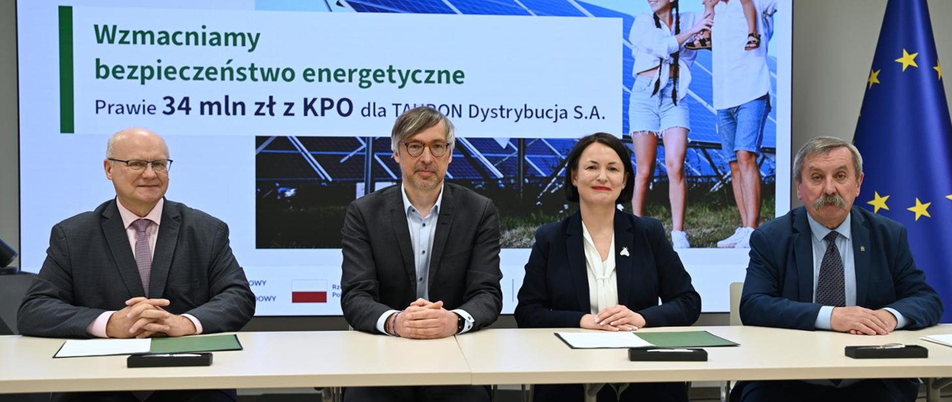 Leszek Kosiorek, Chairman of the Tauron Dystrybucja Management Board, Krzysztof Bolesta, Secretary of State at the Ministry of Climate and Environment, Dorota Zawadzka-Stępniak, President of the Management Board of the NFEP&WM and Józef Matysiak, Vice-President of the Management Board of the NFEP&WM. The above-mentioned individuals are seated next to each other at a table, with folders containing documents and pens placed in front of them. Behind them, a presentation screen is visible, displaying the slogan: “Strengthening energy security. Nearly PLN 34 million from the National Recovery Plan for TAURON Dystrybucja S.A.”.