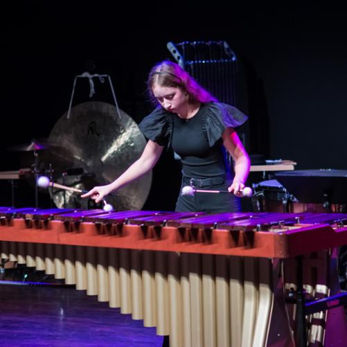 Zdjęcie przedstawia starszą uczestniczkę koncertu grającą czterema pałkami na dużym instrumencie płytkowym perkusyjnym, w tle inne instrumenty perkusyjne