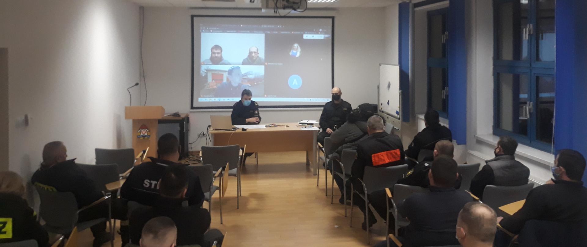 Widok na salę w której znajdują się przedstawiciele Ochotniczych Straży Pożarnych z terenu Powiatu Warszawskiego Zachodniego oraz siedzący przy stole konferencyjnym Komendant i jego Zastępca. Na ekranie widać uczestników wideokonferencji.