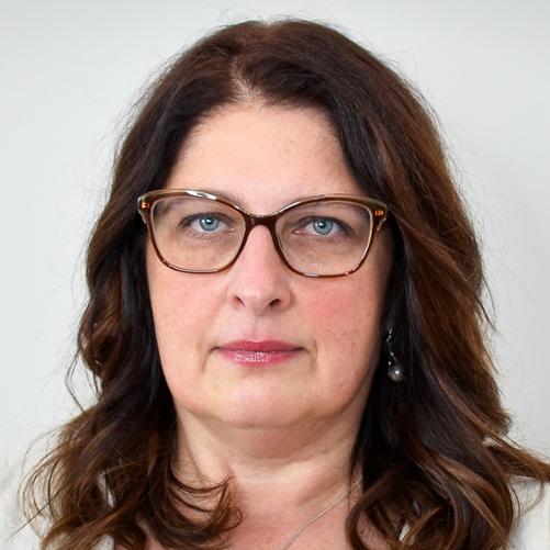 Kobieta w jasnym żakiecie, w okularach, z długimi, rozpuszczonymi ciemnymi włosami w kolorze brązu.