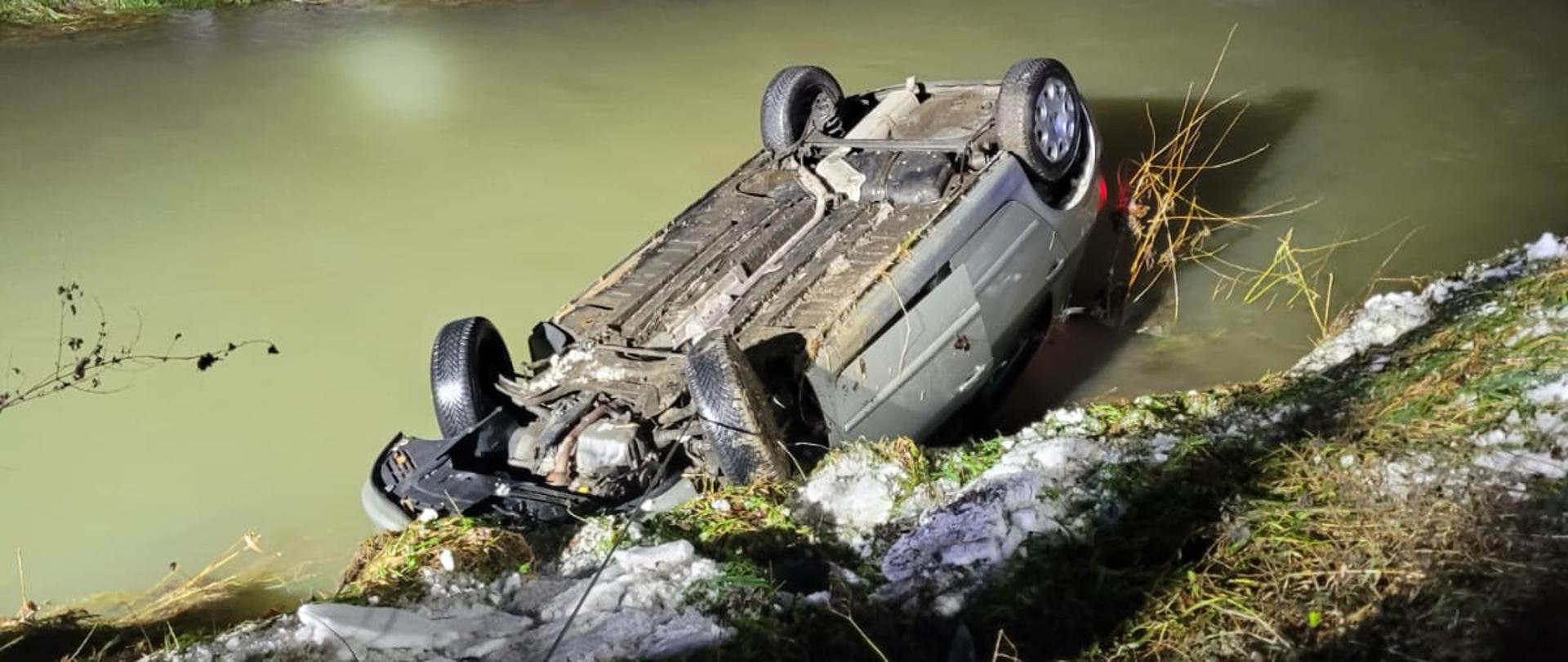 Zdjęcie przedstawia samochód osobowy który wjechał do rzeki. Jest pora nocna. Na zdjęciu widać wywrócony na dachu pojazd osobowy który znajduje się w rzece. Samochód ma skręcone przednie koła i otwarte drzwi od strony kierowcy. Za rzeką znajduje się las. Teren jest oświetlony przez najaśnice z pojazdów pożarniczych.
