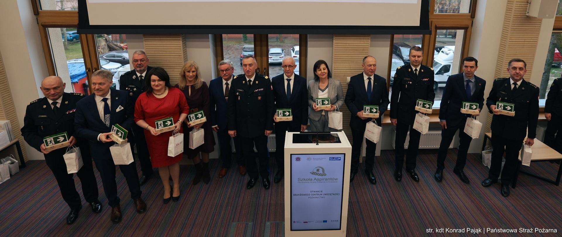 Uczestnicy uroczystości stoją w rzędzie na scenie, trzymając pamiątkowe statuetki, pod ekranem z napisem o otwarciu Branżowego Centrum Umiejętności Pożarnictwo.
