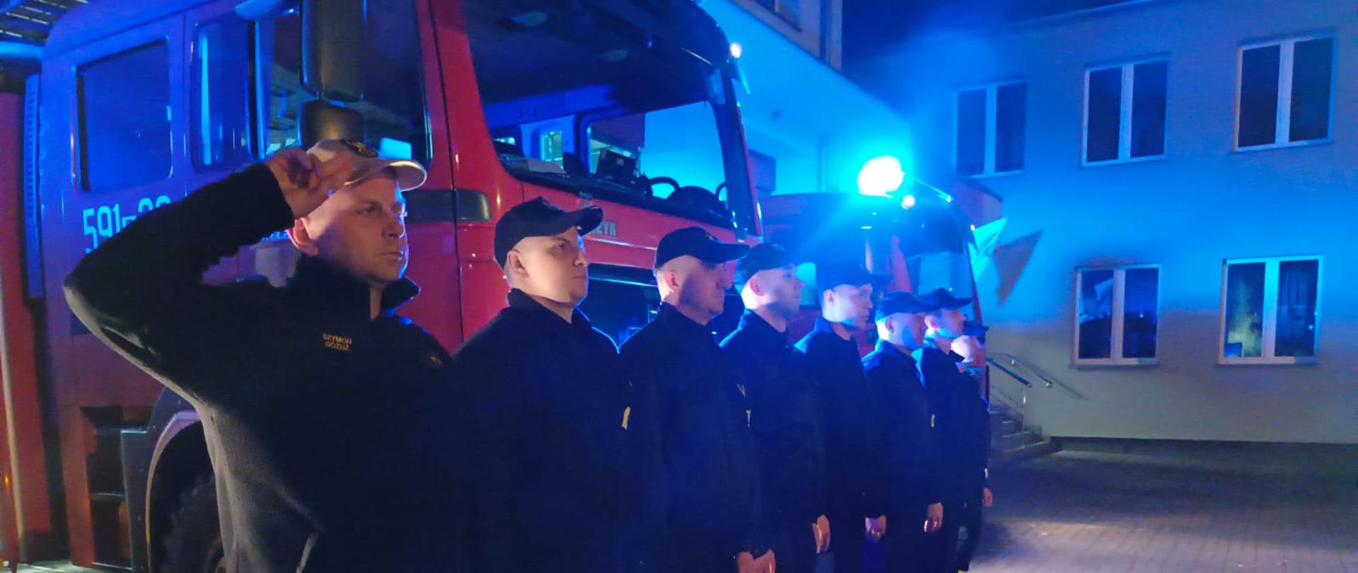 Strażacy oddają hołd, za nimi samochód strażacki, jest ciemno