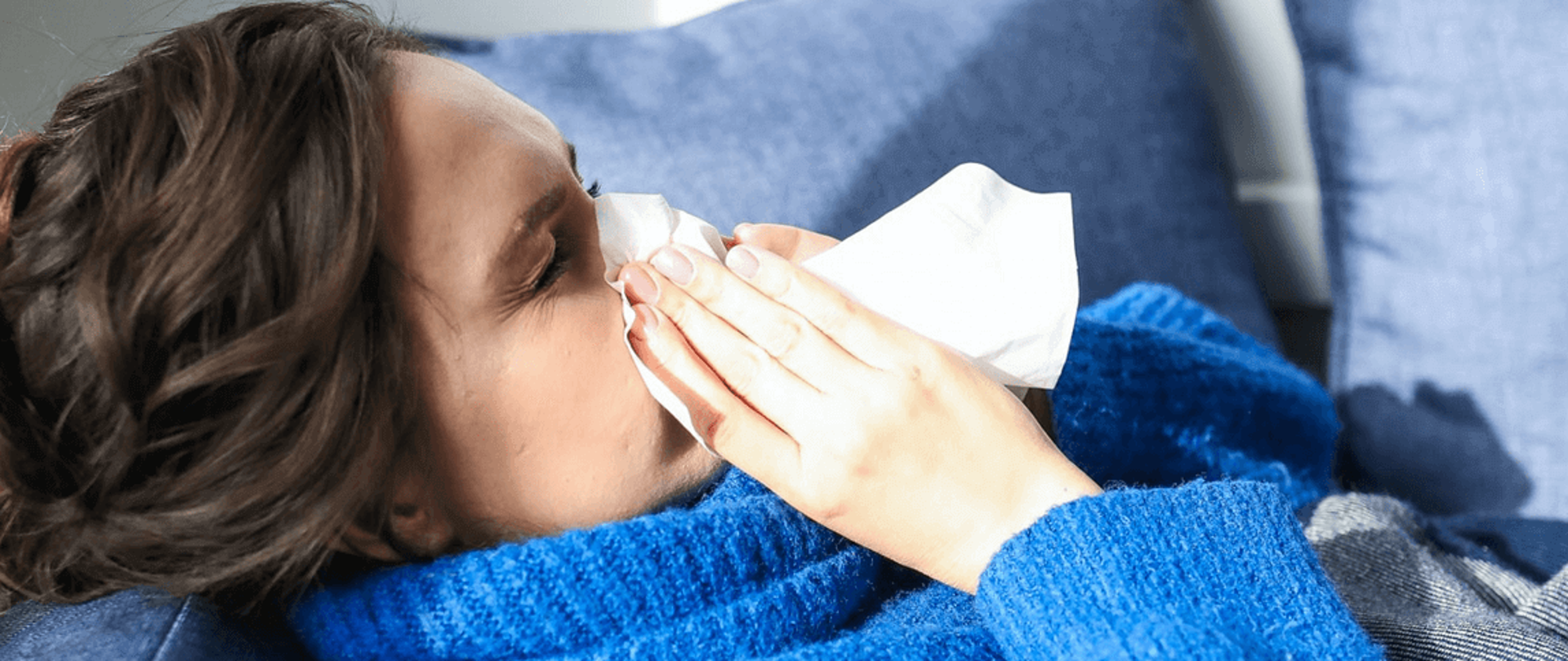 na zdjęciu młoda kobieta w niebieskim swetrze, leżącą na sofie, wycierająca nos w chusteczkę jednorazowego użytku