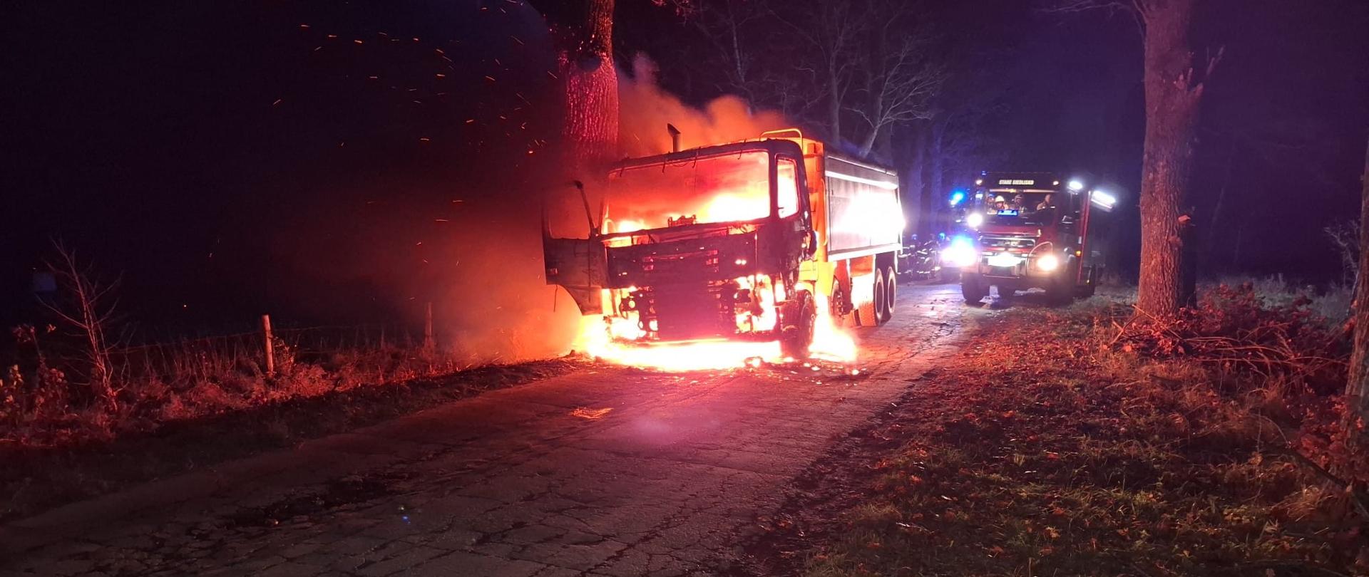 Zdjęcie zrobione nocą. Na drodze płonie ciężarówka, cała w ogniu, duże płomienie. Za nią stoją wozy strażackie. Unosi się dym.