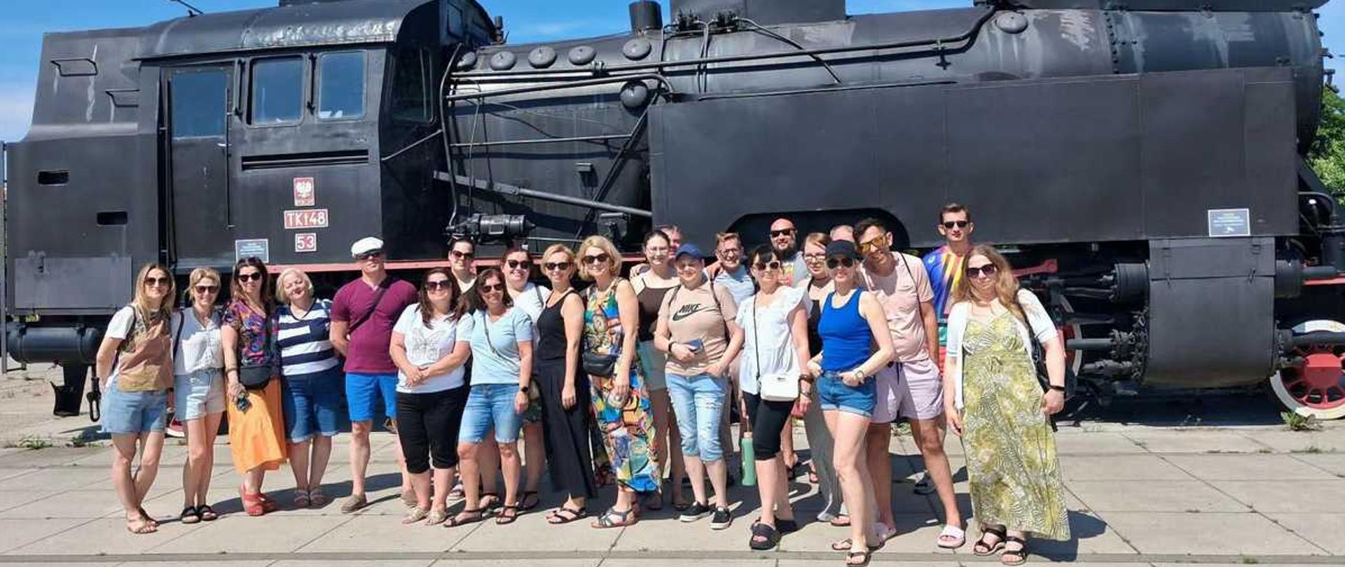Zdjęcie przedstawia grupę dorosłych osób pozujących do wspólnej fotografii na tle zabytkowej, czarnej lokomotywy parowej.LokomotywaWygląd: Wielki, zabytkowy parowóz w kolorze matowej czerni.Oznaczenia: Na boku kabiny maszynisty widnieje czerwona tabliczka z białym napisem TKt48 53 oraz małe godło Polski.Detale: Widoczne są masywne koła z czerwonymi elementami, rury, kotły oraz duży odchylacz dymu z przodu.Ludzie (Grupa)Liczba: Przed lokomotywą stoi obok siebie około 20 osób.Ubiór: Wszyscy są ubrani w letnie, codzienne stroje – krótkie spodenki, koszulki, spódnice, sukienki i sandały lub klapki. Większość osób ma na sobie okulary przeciwsłoneczne.Nastroje: Ludzie są uśmiechnięci, stoją blisko siebie w luźnej, wycieczkowej atmosferze.Otoczenie i tłoPodłoże: Grupa stoi na płaskim placu ułożonym z dużych, betonowych płyt.Pogoda: Scena ma miejsce w pełnym, jasnym słońcu.Niebo: Tło nad lokomotywą stanowi całkowicie bezchmurne, jasnoniebieskie niebo.