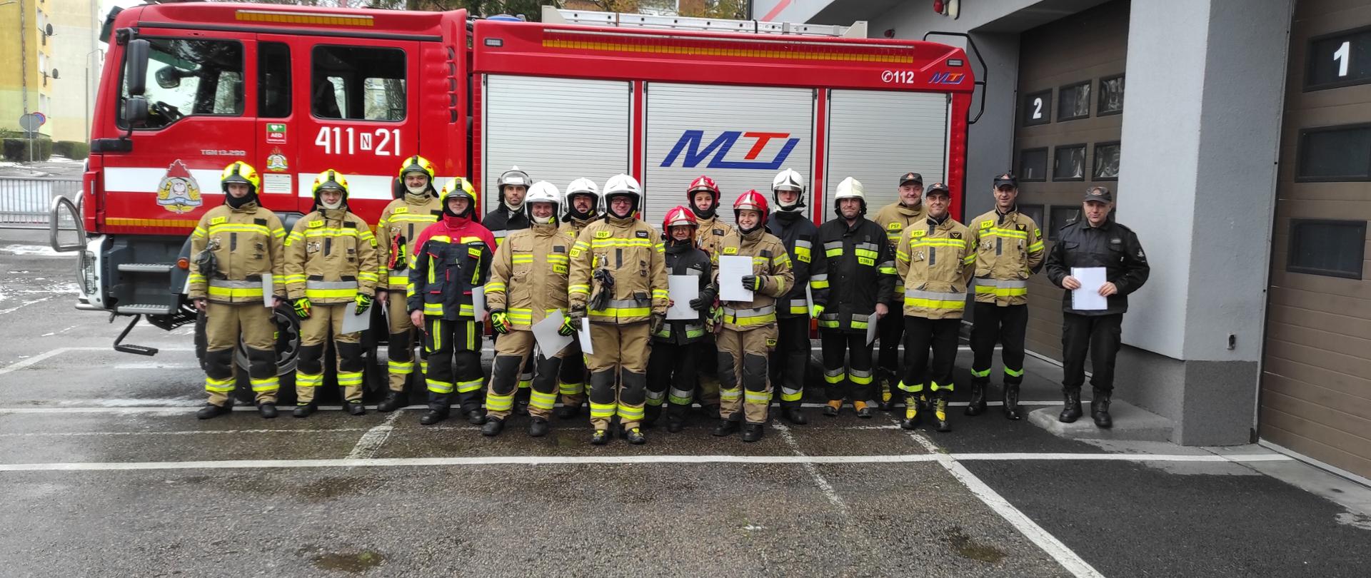 Na tle dużego czerwonego samochodu strażackiego stoją ustawieni obok siebie strażacy w mundurach bojowych i hełmach. Z prawej strony budynek strażnicy.
