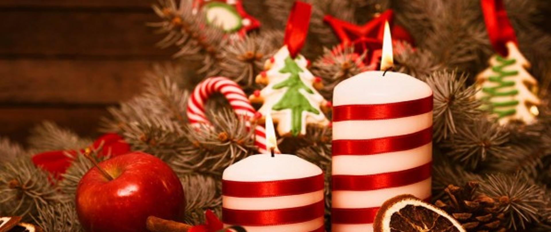 zdjęcia w czerwono ciepłych odcieniach przedstawia stroik świąteczny, który zawiera pierniczki, dwie świeczki, jabłko, igliwie i laski cukrowe