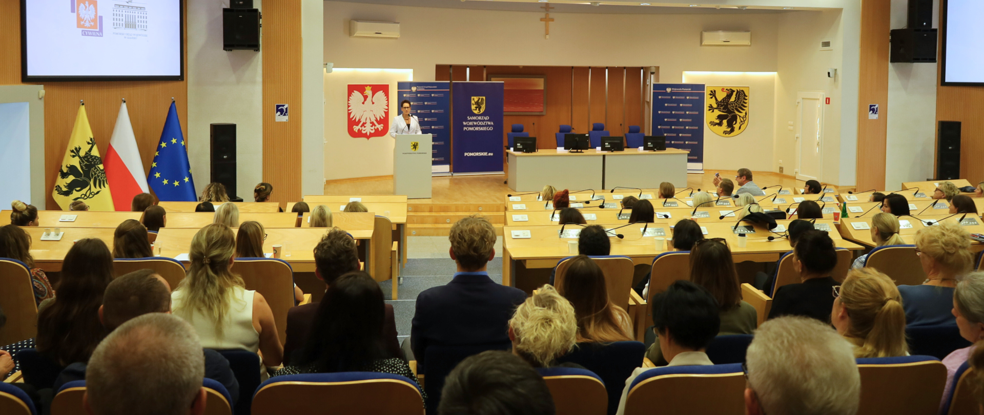 duża sala, w niej ludzie słuchający wykładu osoby stojącej przy mównicy