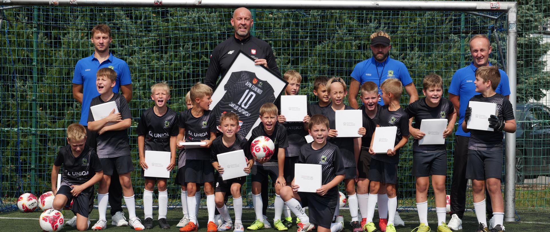 Zdjęcie zbiorowe. Minister sportu i turystyki pozuje do zdjęcia z trenerami i zawodnikami klubu APN Funinio. Zdjęcie zrobione na boisku piłkarskim w bramce. Dzieci trzymają w rękach podarunki-pudełka z logotypem MSiT. Minister trzyma oprawioną w ramę czarną koszulkę z nazwą klubu numerem 10 i swoim imieniem i nazwiskiem. Na ziemi leżą piłki.