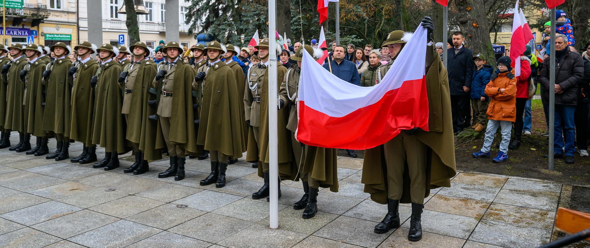 Żołnierze w mundurach stoją w szyku na Placu Wolności w Rzeszowie podczas podnoszenia flagi państwowej, w otoczeniu licznie zgromadzonych mieszkańców.