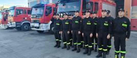 Strażący stoją na baczność w dwuszeregu na tle samochodów strażackich.