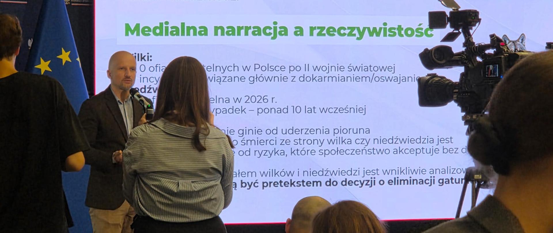 Mikołaj Dorożała - Wiceminister Klimatu i Środowiska, Główny Konserwator Przyrody udziela wywiadu mediom, w tle widać prezentację o tytule " Medialna narracja a rzeczywistość", w prawym górnym rogu widać kamerę, a na pierwszym planie osoby stojące i siedzące.
