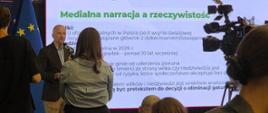 Mikołaj Dorożała - Wiceminister Klimatu i Środowiska, Główny Konserwator Przyrody udziela wywiadu mediom, w tle widać prezentację o tytule " Medialna narracja a rzeczywistość", w prawym górnym rogu widać kamerę, a na pierwszym planie osoby stojące i siedzące.