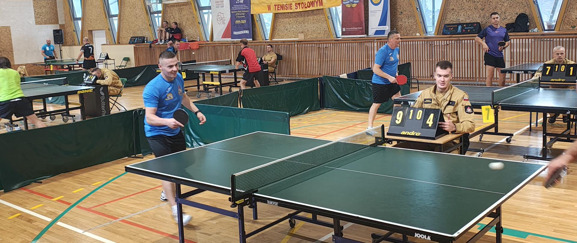 Ujęcie kilku stołów do tenisa w trakcie gier. Na pierwszym planie trwa wymiana piłki, obok sędzia w beżowym mundurze obsługuje tablicę wyników. W tle banery mistrzostw i inni zawodnicy.
Tekst na banerze (w tle): „Mistrzostwa Województwa Podkarpackiego Strażaków PSP w tenisie stołowym”.