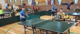Ujęcie kilku stołów do tenisa w trakcie gier. Na pierwszym planie trwa wymiana piłki, obok sędzia w beżowym mundurze obsługuje tablicę wyników. W tle banery mistrzostw i inni zawodnicy.