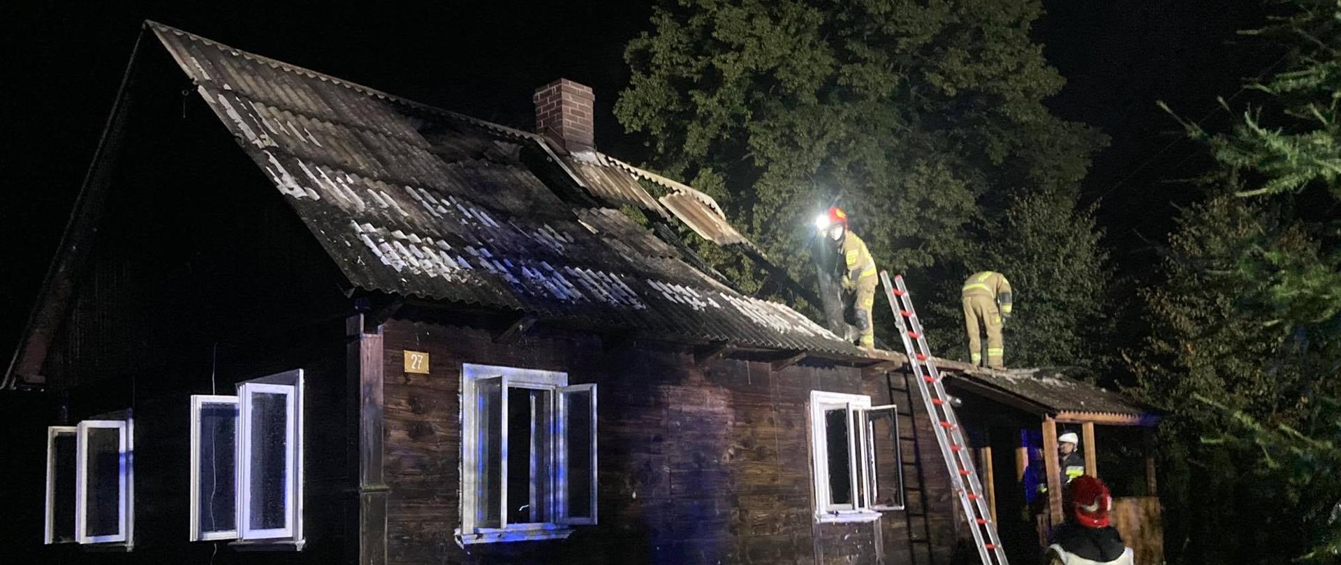 Drewniany budynek mieszkalny po pożarze. Część konstrukcji dachu zawalona. Na dachu oraz przed budynkiem pracujący strażacy.