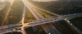 Węzeł Zabrze Południe na A4 o poranku. Promienie słońca ogrzewają i oświetlają infrastrukturę drogową, w tym wiadukt nad autostradą i linią kolejową. Na fotografii ruch pojazdów jest niewielki.